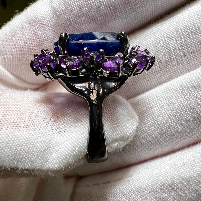 May include: A dark metal ring featuring a large, rectangular, faceted blue gemstone surrounded by smaller, round, purple gemstones. The ring's band is a simple, solid design.
