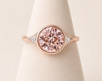 May include: A rose gold ring featuring a large, round, peach-colored center stone set in a bezel. The band includes two small, triangular-cut clear stones on either side of the central setting, creating a balanced and symmetrical design.