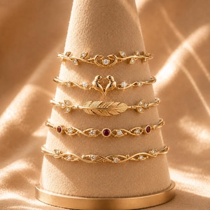 Pode incluir: Um conjunto de cinco pulseiras douradas empilhadas num expositor cónico. Os designs incluem um coração com uma pedra central, dois pássaros frente a frente, uma pena texturizada, uma corrente com pedras vermelhas e claras e uma banda torcida.