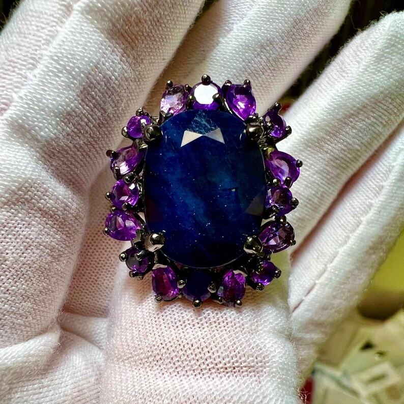 May include: A large, oval-shaped, dark blue gemstone ring, surrounded by smaller purple gemstones. The ring has a dark metal setting. The gemstones are set against a white background.