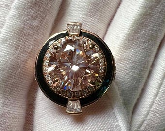 May include: A rose gold ring with a large, round, light brown gemstone at the center, surrounded by smaller clear stones. The ring has a black enamel band and rectangular clear stones on the top and bottom.