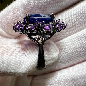 May include: A dark metal ring featuring a large, rectangular, faceted blue gemstone surrounded by smaller, round, purple gemstones. The ring's band is a simple, solid design.
