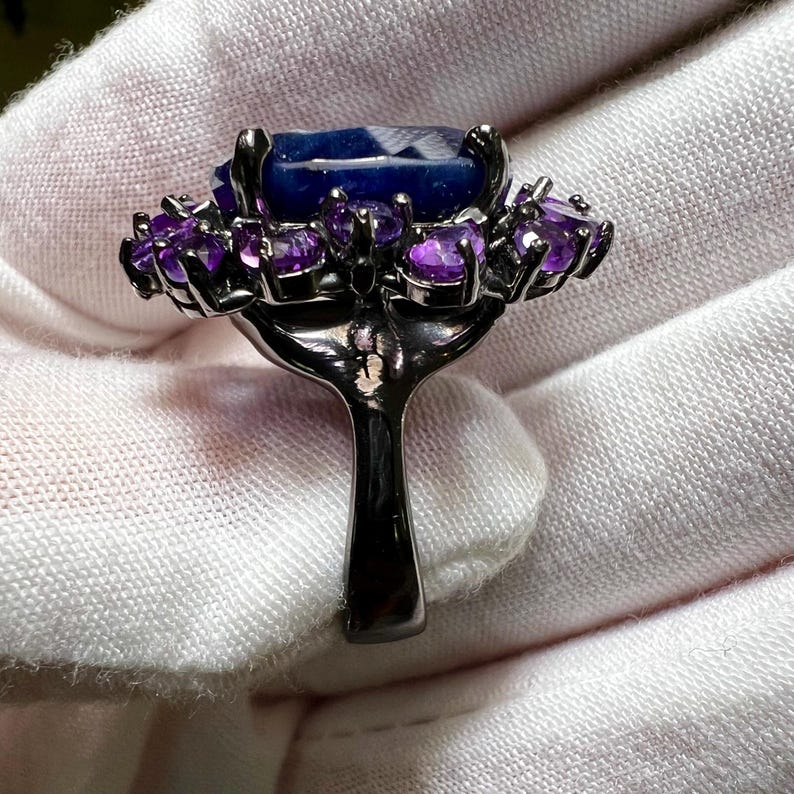 May include: A dark metal ring featuring a large, rectangular, faceted blue gemstone surrounded by smaller, round, purple gemstones. The ring's band is a simple, solid design.