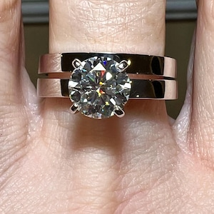 Pode incluir: Um close-up de um anel de noivado de prata com um grande diamante redondo e transparente. O anel é usado no dedo de uma pessoa.