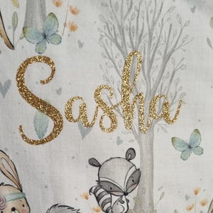 Könnte beinhalten: Ein weißer Stoff mit Waldmotiv, auf dem der Name "Sasha" in goldenen Glitzerbuchstaben und Illustrationen von Tieren, Bäumen und Schmetterlingen zu sehen ist.