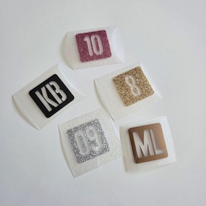 Peut inclure: Cinq transferts thermocollants carrés présentant des motifs et textures variés. L'un affiche le nombre 10 en paillettes rose foncé, le 8 en paillettes dorées, les lettres KB en noir, le nombre 09 en paillettes argentées et les lettres ML en bronze.