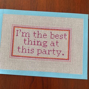 May include: A needlepoint canvas with the text "I'm the best thing at this party." The text is in purple, framed by an orange and purple border, and set against a white background. The canvas is bordered by light blue tape.