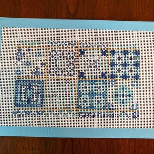 May include: A hand-stitched needlepoint canvas featuring a geometric design in shades of blue and teal. The design is divided into six squares with intricate patterns, framed by a light blue border. The canvas is on a white background.