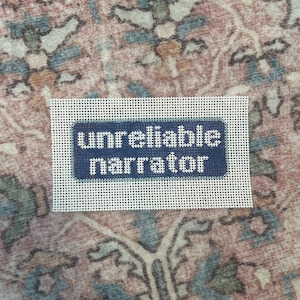 May include: A needlepoint canvas with the words "unreliable narrator" stitched in white on a navy blue background. The canvas is set against a patterned, muted pink and blue rug.