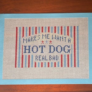 May include: A rectangular needlepoint canvas featuring a light blue border and a central design of vertical red, white, and blue stripes. The text reads: MAKES ME WANT A HOT DOG REAL BAD, stitched in dark blue letters with small red and blue accents.