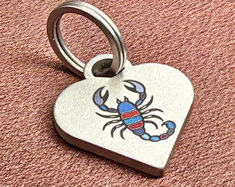 May include: A heart-shaped metal tag featuring a colorful scorpion graphic with blue, red, and purple enamel details. The tag has a brushed silver finish and includes a circular metal split ring attached to the top for secure fastening.