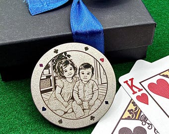 May include: A circular silver-toned metal poker chip featuring a laser-engraved portrait of two children. The chip is decorated with small card suit symbols around the edge and is positioned next to a black gift box with a blue ribbon and playing cards.