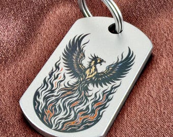 May include: A silver-toned metal dog tag keychain featuring a detailed black and orange graphic of a phoenix rising from flames. The rectangular tag has rounded corners and a circular metal split ring attached at the top center.