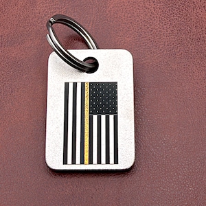 May include: A rectangular silver-toned metal keychain tag featuring a printed graphic of a black and white American flag with a single vertical gold-colored stripe. The tag includes a circular hole at the top with a dark metal split ring attached.