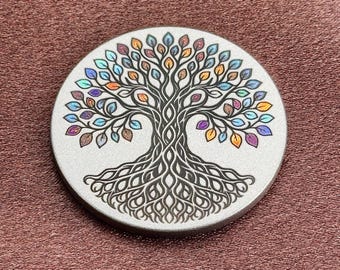 May include: A circular silver-toned metal disc featuring a detailed black engraved design of a tree with intricate roots and branches. The leaves are filled with a variety of iridescent, multi-colored hues including blue, purple, orange, and green.