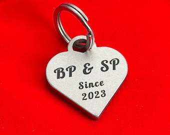May include: A heart-shaped silver-toned metal keychain tag featuring a split ring at the top. The surface is engraved with the black text BP & SP followed by the word Since and the year 2023 centered below the initials.