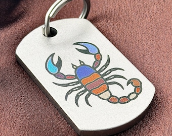 May include: A rectangular silver-toned metal tag with rounded corners featuring a colorful scorpion graphic. The scorpion is depicted with blue, purple, and orange segments and black outlines. A circular metal split ring is attached to the top hole.