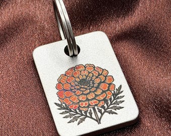 May include: A rectangular silver-toned metal keychain tag featuring a detailed, engraved illustration of a vibrant orange marigold flower with green leaves. The tag has a smooth, brushed finish and includes a circular metal split ring at the top.