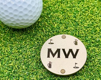 May include: A white dimpled golf ball positioned next to a circular metallic golf ball marker. The marker features the letters MW in the center, surrounded by small icons of golf flags, golf bags, and golf balls engraved on the surface.