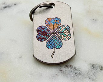 May include: A rectangular metal keychain tag with a matte grey finish featuring a laser-engraved four-leaf clover design. The clover is filled with intricate, iridescent patterns in shades of blue, purple, orange, and gold. A dark metal ring is attached.