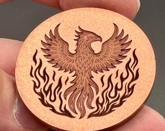 May include: A circular copper-colored metal coin featuring a deeply engraved design of a phoenix with wings spread wide, surrounded by stylized flames. The surface has a textured, metallic finish with dark recessed lines.