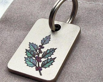 May include: A rectangular metal keychain tag with a brushed finish featuring a detailed illustration of a holly branch with green leaves and red berries. The tag has a circular hole at the top attached to a standard silver-toned metal split ring.
