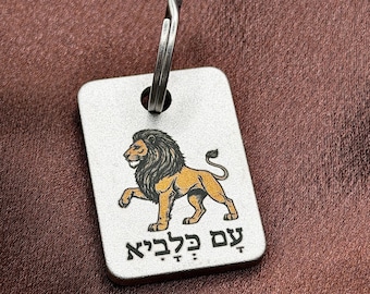 May include: A rectangular silver-toned metal tag featuring a colored illustration of a lion in profile. Below the lion, the Hebrew text "עַם כְּלָבִיא" is engraved in black. A metal split ring is attached through a hole at the top of the tag.