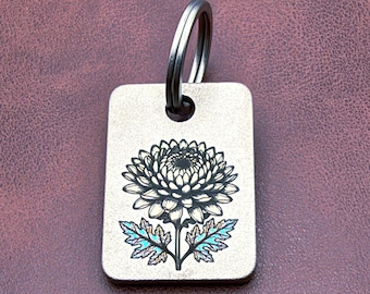 May include: A rectangular metal keychain tag with a brushed silver finish featuring a black engraved illustration of a chrysanthemum flower. The leaves at the base of the flower have a subtle iridescent blue and green tint. A metal split ring is attached.
