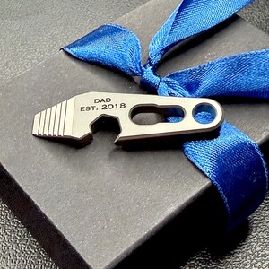 May include: A metallic, multi-functional tool featuring a bottle opener and a serrated edge, engraved with the text DAD EST. 2018. The tool is positioned on a black box adorned with a vibrant blue ribbon tied in a bow across the top surface.