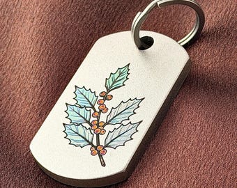 May include: A rectangular silver-toned metal tag with rounded corners and a circular split ring attached at the top. The surface features a detailed illustration of a holly branch with light blue leaves and small orange berries.