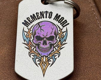 May include: A silver-toned metal dog tag keychain featuring a purple skull graphic with a cracked forehead. The text MEMENTO MORI is printed in black above the skull, which is surrounded by orange and blue tribal-style decorative accents.