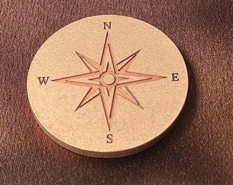 May include: A circular copper-toned metal coin featuring an engraved compass rose design. The center displays a star-shaped directional marker with the letters N, S, E, and W positioned at the top, bottom, right, and left points respectively.