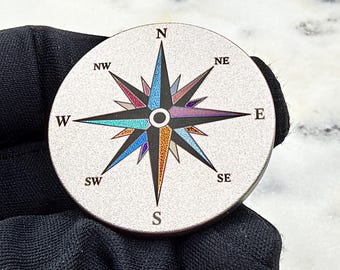 May include: A circular metal disc featuring a detailed compass rose design. The compass points are rendered in vibrant, iridescent colors including blue, purple, and orange, set against a textured silver background with black cardinal direction letters.