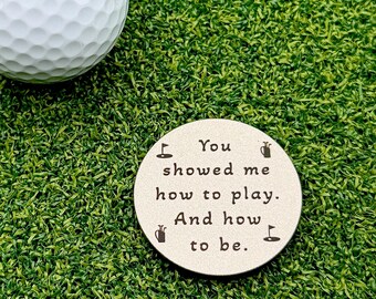 May include: A white dimpled golf ball and a round, light-colored metal golf ball marker engraved with the text: You showed me how to play. And how to be. The marker features small black icons of golf flags and golf bags in the corners.