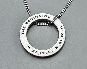 May include: A silver-toned circular pendant necklace featuring a thin metal chain. The flat, ring-shaped charm is engraved with the text THE BEGINNING and the coordinates 41° 04' 15" N 71° 51' 25" W in a clean, black, sans-serif font.