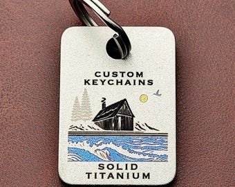 May include: A rectangular solid titanium keychain with a split ring. The surface features the text CUSTOM KEYCHAINS and SOLID TITANIUM, with a graphic illustration of a cabin, pine trees, a yellow sun, a bird, and blue ocean waves.