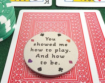 May include: A round, silver-toned metal token featuring the black engraved text, "You showed me how to play. And how to be." The token is decorated with small, colorful card suit symbols including clubs, spades, hearts, and diamonds.
