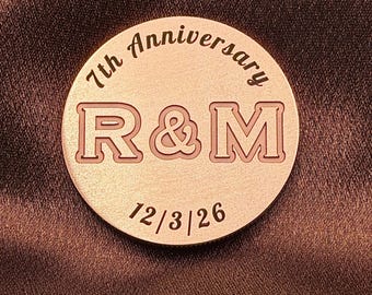 May include: A circular copper-toned metal token featuring the engraved text 7th Anniversary at the top, the letters R & M in the center, and the date 12/3/26 at the bottom. The surface has a brushed metallic finish with dark etched lettering.