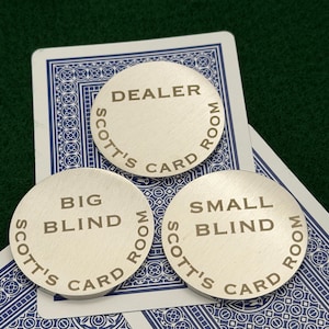 May include: Three silver poker chips with the text "DEALER", "BIG BLIND", and "SMALL BLIND". Each chip has "SCOTT'S CARD ROOM" around the edge. The chips are on top of playing cards.