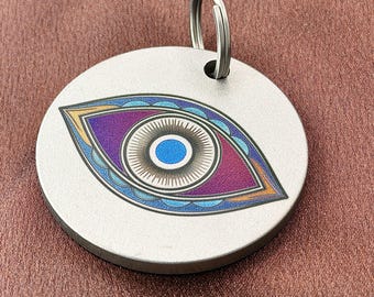 May include: A circular silver-toned metal tag featuring a stylized eye graphic in shades of blue, purple, and orange. The eye design includes a central blue pupil, a detailed iris, and decorative outer layers. A metal split ring is attached at the top.