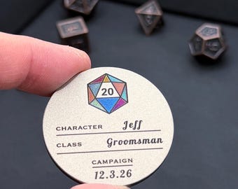 May include: A circular metal token with a matte finish featuring a colorful 20-sided die graphic at the top. The text reads CHARACTER Jeff, CLASS Groomsman, CAMPAIGN 12.3.26. Several dark copper-colored polyhedral dice are visible in the background.