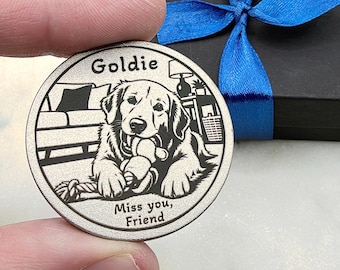May include: A circular silver-toned metal token featuring an engraved illustration of a Golden Retriever dog holding a rope toy. The text Goldie is at the top and Miss you, Friend is at the bottom. The background shows a sofa and a lamp.