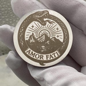 May include: A round, silver-colored metal coin with an engraved design. The design features a snake eating its tail, surrounding a mountain scene with a rising sun and the words "AMOR FATI". The coin is held by a gloved hand.