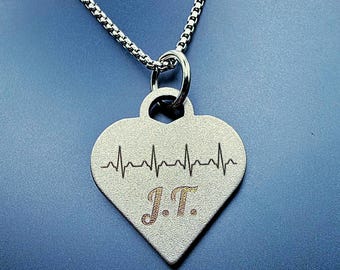 May include: A silver-toned heart-shaped pendant necklace featuring an engraved black heartbeat line across the top and the letters J.T. in a cursive font below. The pendant hangs from a matching silver-toned box chain link necklace.