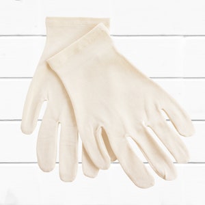 Puede incluir: Un par de guantes de color marfil, probablemente hechos de una tela suave y elástica. Los guantes se muestran planos, con los dedos ligeramente separados. El fondo es una superficie blanca con listones horizontales.