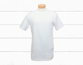 Può includere: Una maglietta bianca a maniche corte, esposta su un manichino. La maglietta ha uno scollo tondo e un design semplice e classico. Lo sfondo è un muro di legno bianco.