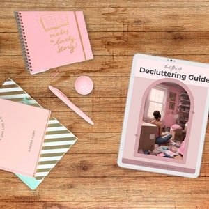 May include: A pink tablet computer displaying a decluttering guide with the text "Decluttering Guide" and "thel t" at the top. The guide features a photo of a woman sitting in a messy room, surrounded by boxes and clothes. The text "www.yourwebsite.com" is at the bottom of the guide.