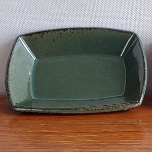 Może przedstawiać: Dwie prostokątne ceramiczne miseczki z lekko zakrzywionymi bokami i zaokrąglonymi rogami. Miseczka po lewej stronie ma ciemnozieloną glazurę z teksturowanym, nakrapianym brzegiem, podczas gdy ta po prawej ma gładkie, błyszczące turkusowe wykończenie.