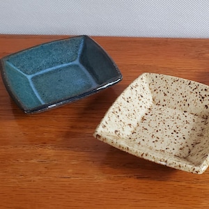May include: Three small, square ceramic dishes in shades of teal, speckled beige, and brown. The dishes have a slightly raised rim and are displayed on a wooden surface. These decorative dishes are suitable for serving small portions.