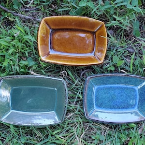 May include: Three rectangular ceramic dishes with raised edges. One dish is a glossy amber brown, one is a muted sage green, and one is a vibrant speckled blue. Each dish features a smooth, glazed finish and a slightly curved, shallow basin design.
